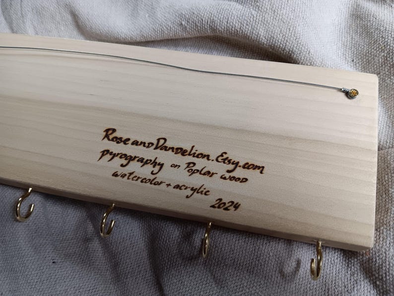 May include: Wooden key holder with five hooks and a burned-in inscription: "Rose and Dandelion.Etsy.com Pyrography on Poplar wood Watercolor + acrylic 2024"