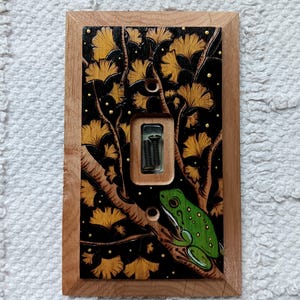Puede incluir: Una placa de interruptor de luz rectangular de madera con un diseño pintado a mano de una rana verde sobre una rama entre hojas de ginkgo doradas sobre un fondo negro. La placa incluye un recorte central rectangular y dos orificios para tornillos.