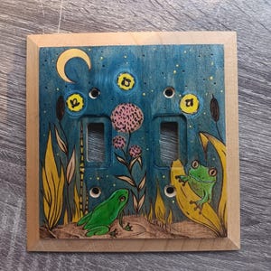Puede incluir: Cubierta de interruptor de luz doble de madera con un diseño caprichoso pintado a mano. La obra de arte presenta dos ranas verdes, plantas, una luna creciente y detalles amarillos sobre un fondo azul. La cubierta está enmarcada con un borde de madera natural.