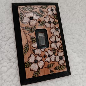 May include: A rectangular wooden light switch plate featuring a black border and a natural wood center decorated with a pyrography design of white dogwood flowers and green leaves. The center includes a black toggle switch cutout with two screws.