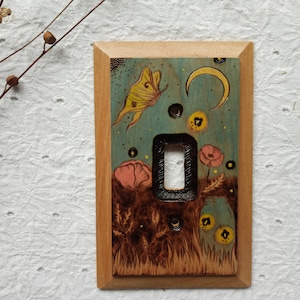 May include: Wooden light switch cover with a hand-painted design. The artwork features a yellow butterfly, a crescent moon, and pink poppies against a teal background. The switch plate is framed in natural wood.