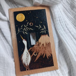 May include: Wooden light switch cover featuring a hand-painted design. The artwork depicts a crane with a mountain, moon, and stars against a dark background. The frame is a light wood color, and the design is detailed with various colors.