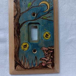 May include: A wooden light switch cover with a hand-painted design of a night scene with a crescent moon, stars, a tree, mushrooms, and glowing orbs.