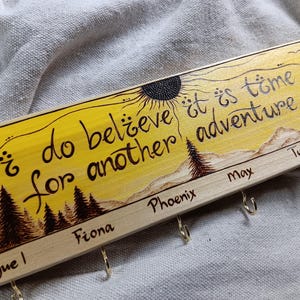 May include: A wooden key holder with a yellow background and a sun design. The text "I do believe it is time for another adventure" is written in black. There are five hooks labelled "Rogue I", "Fiona", "Phoenix", "Max", and "Turtle".