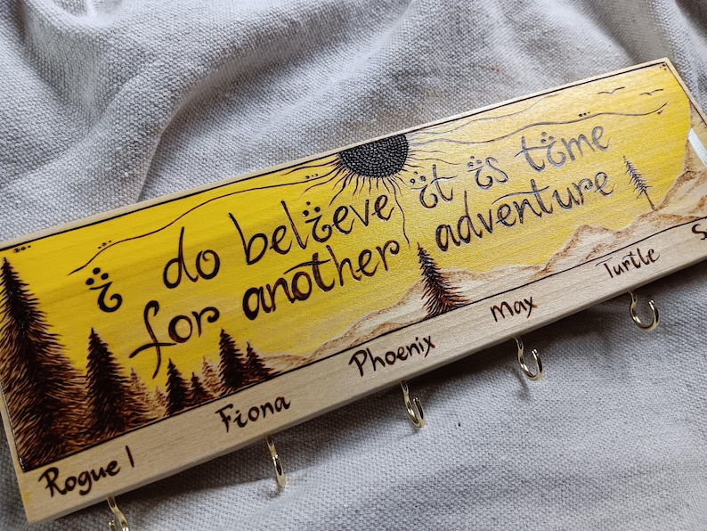 May include: A wooden key holder with a yellow background and a sun design. The text "I do believe it is time for another adventure" is written in black. There are five hooks labelled "Rogue I", "Fiona", "Phoenix", "Max", and "Turtle".