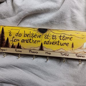 May include: Wooden key holder with a yellow background and a burnt wood design. The text reads "&iquest;do believe it is time for another adventure". There are six hooks labelled "Rogue", "Fiona", "Phoenix", "Max", "Turtle", and "Spares".