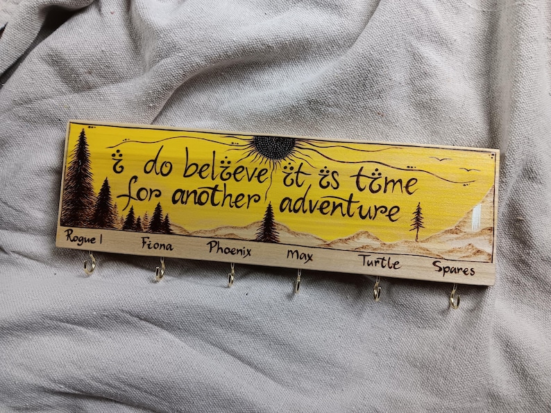 May include: Wooden key holder with a yellow background and a burnt wood design. The text reads "&iquest;do believe it is time for another adventure". There are six hooks labelled "Rogue", "Fiona", "Phoenix", "Max", "Turtle", and "Spares".