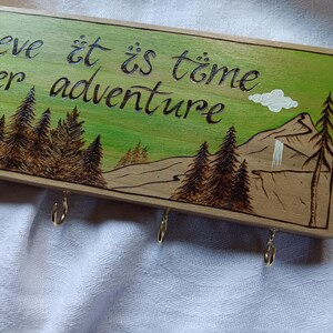 May include: Wooden key holder with a green background and a mountain scene. The text "Believe it is time for another adventure" is written in black cursive. There are five metal hooks on the bottom.