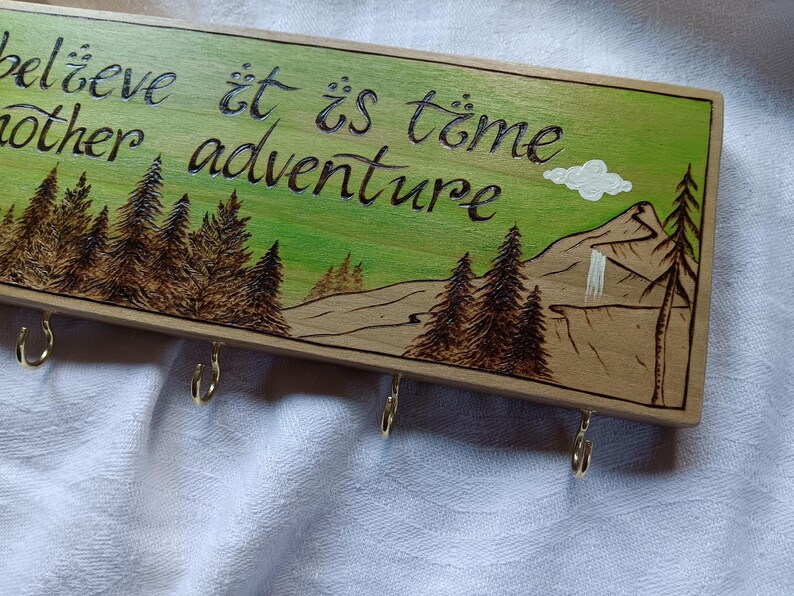 May include: Wooden key holder with a green background and a mountain scene. The text "Believe it is time for another adventure" is written in black cursive. There are five metal hooks on the bottom.