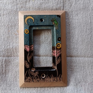 May include: A rectangular wooden light switch cover plate featuring a hand-painted design of a yellow crescent moon, a small green creature, and pink flowers against a dark blue and black background, framed by a natural light-colored wood border.