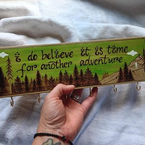 May include: A wooden key holder with a green background and a burnt wood design. It features a quote that reads "I do believe it is time for another adventure." There are five hooks on the bottom of the key holder.