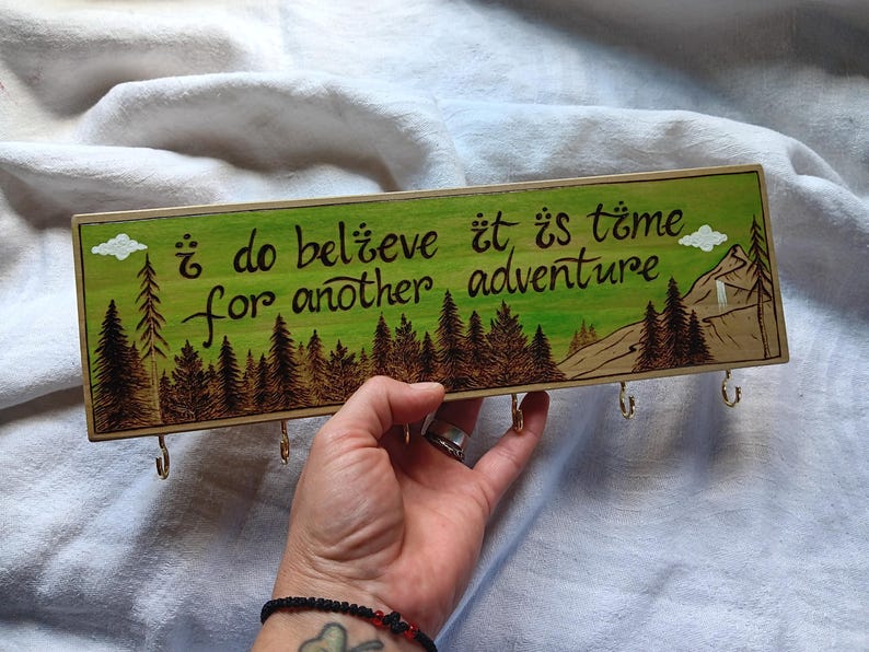 May include: A wooden key holder with a green background and a burnt wood design. It features a quote that reads "I do believe it is time for another adventure." There are five hooks on the bottom of the key holder.