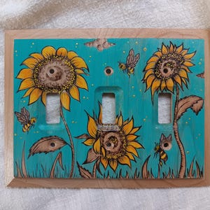 Puede incluir: Una cubierta de interruptor triple de madera pintada a mano con girasoles y abejas sobre un fondo turquesa. Los girasoles son amarillos y marrones, y las abejas son amarillas y negras. La cubierta tiene un marco de madera natural.