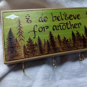 May include: Wooden key holder with a green background and a wood-burned design of a forest with the text "i do believe it is for another adventure".