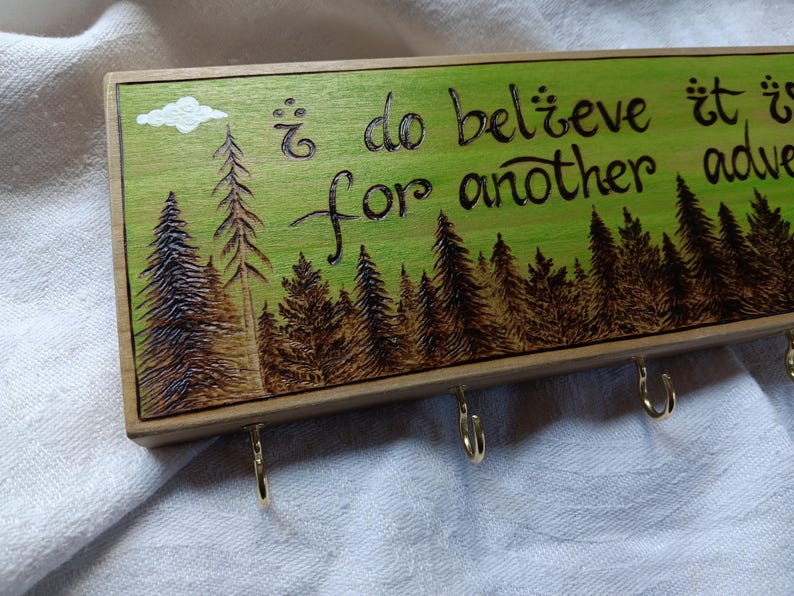 May include: Wooden key holder with a green background and a wood-burned design of a forest with the text "i do believe it is for another adventure".