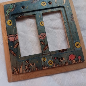 May include: A rectangular wooden double light switch plate featuring a dark teal stained finish. The surface is decorated with hand-painted illustrations of pink flowers, red mushrooms, yellow circular motifs, and a crescent moon against a dark background.