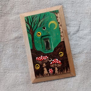 Puede incluir: Una tapa de interruptor de luz de madera con un diseño pintado a mano de una escena de bosque con setas, una luna creciente y un interruptor de luz. El fondo es verde con estrellas amarillas y las setas son rojas con manchas blancas.