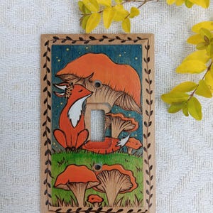 May include: A rectangular wooden light switch plate featuring a hand-painted scene of an orange fox sitting in a green field with orange mushrooms. The design includes a dark blue sky with yellow dots and a decorative leaf border burned into the wood.