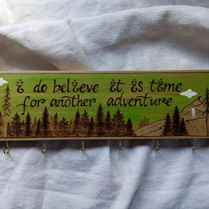 May include: Wooden key holder with a green background and a burnt wood design. It features a quote that reads "I do believe it is time for another adventure".