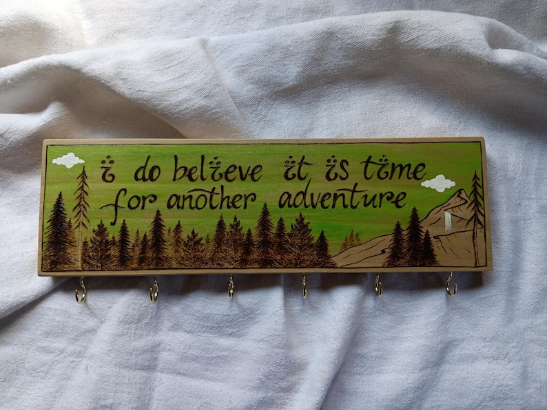 May include: Wooden key holder with a green background and a burnt wood design. It features a quote that reads "I do believe it is time for another adventure".