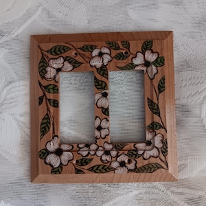 May include: A rectangular wooden double light switch plate featuring a hand-painted design of white dogwood flowers with dark outlines and green leaves. The light-toned wood grain is visible throughout the frame, which includes two rectangular cutouts.
