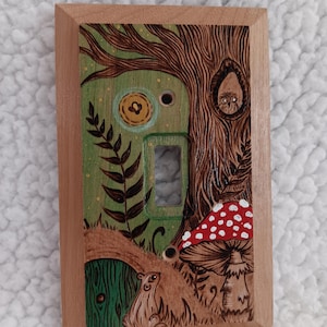 May include: A wooden light switch cover featuring a whimsical forest scene. The design includes a tree with an owl, a mushroom with red and white spots, and a small green door. The background is green with a light brown wooden frame.