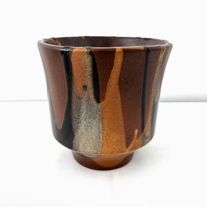 Op de afbeelding: Een keramische beker met een uniek, gedruipt glazuurontwerp. De beker heeft een kleurenpalet van bruin, zwart en goud. De beker heeft een uitlopende rand en een voet. De beker is waarschijnlijk handgemaakt en geschikt voor dranken.