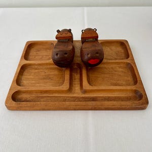 Puede incluir: Bandeja de madera para servir con cuatro compartimentos y un divisor central. Dos figuras de hipopótamos de madera tallada se asientan en la bandeja. Un hipopótamo tiene la boca pintada de rojo. La bandeja y los hipopótamos son de madera marrón cálido.
