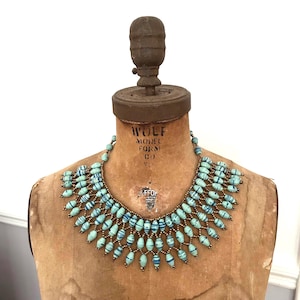 May include: A turquoise beaded necklace with blue striped accents. The necklace is displayed on a vintage mannequin. The beads are strung together with a bronze-coloured wire, creating a unique and eye-catching design.