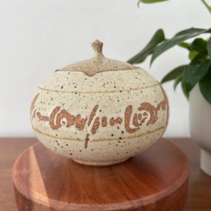 May include: A speckled, beige ceramic jar with a lid. The jar has a rounded shape and is decorated with brown, stylized lettering. The lid has a small, pointed finial. The jar sits on a round, wooden base.