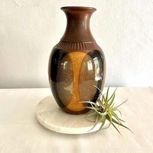 Op de afbeelding: Een bruine keramische vaas met een uniek ontwerp. De vaas heeft een donkerbruine bovenkant en een body met zwarte, gouden en bruine geometrische vormen. De vaas staat op een witte marmeren schaal met een kleine plant.