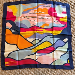May include: Square silk scarf with a colorful abstract landscape design. The scarf features a navy blue border and a vibrant palette of pink, orange, yellow, and blue. The scarf has the text "Liberty" and "Made in England".