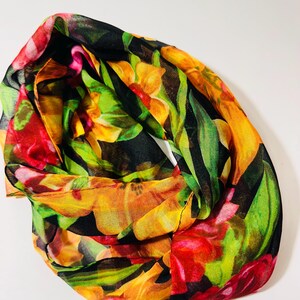 May include: A colorful floral scarf with a black background. The scarf features vibrant flowers in shades of red, yellow, and green. The scarf is made of a lightweight, sheer fabric, and is folded in a circular shape.