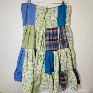 May include: A patchwork skirt with a variety of fabrics and colors. The skirt features blue, green, and floral patterned panels. The skirt is hanging on a metal hanger against a white background.
