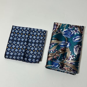 May include: Two folded pocket squares. One is navy blue with a repeating pattern of small squares and rectangles in shades of blue, yellow, and white. The other is teal with a floral pattern of blue and purple tulips and brown leaves.