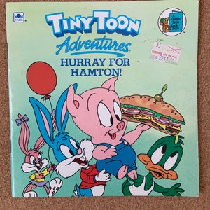 Peut inclure: Une couverture de livre mettant en scène les personnages de Tiny Toon Adventures, dont Buster Bunny, Hamton Pig et Plucky Duck. Le titre du livre est "Hurray for Hamton!" et fait partie de la série Golden Look-Look Book.