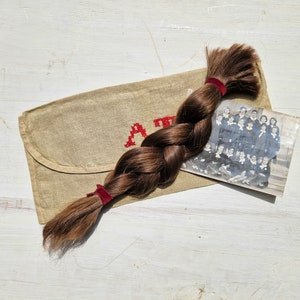 Puede incluir: Una larga trenza marrón de cabello está envuelta alrededor de un sobre de tela beige con costuras rojas que dicen "Un Recuerdo". Una foto en blanco y negro de un grupo de personas está metida dentro del sobre.