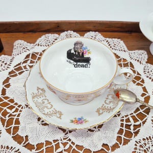 May include: A white teacup and saucer set with gold trim and floral accents. The teacup features a graphic of a person at a piano with the text "who's dead?" A silver spoon rests on the saucer.
