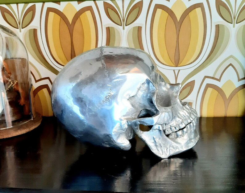 May include: A silver skull sculpture with a smooth, shiny finish. The skull is positioned on a dark surface with a patterned background.