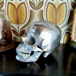 May include: A silver skull sculpture with a detailed design. The skull is sitting on a dark surface with a patterned background.