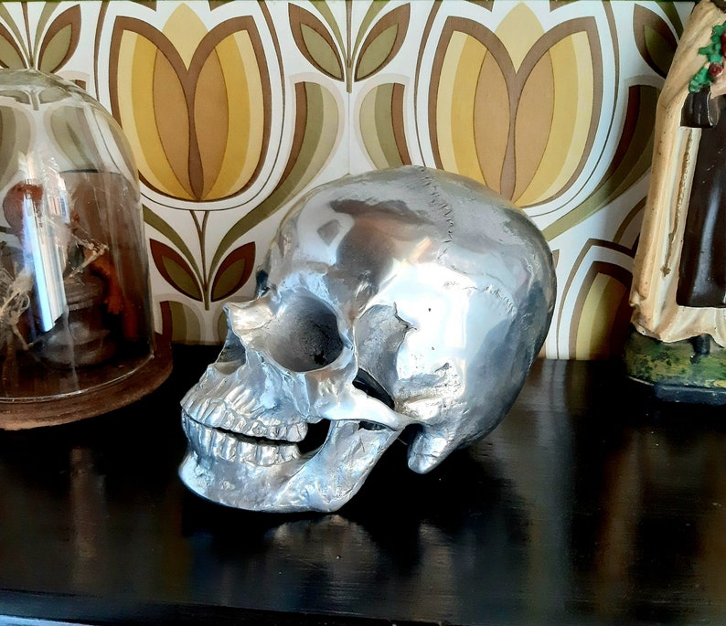May include: A silver skull sculpture with a detailed design. The skull is sitting on a dark surface with a patterned background.