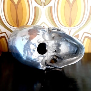 May include: A silver skull sculpture with a detailed design. The skull is hollow and has a large eye socket opening.
