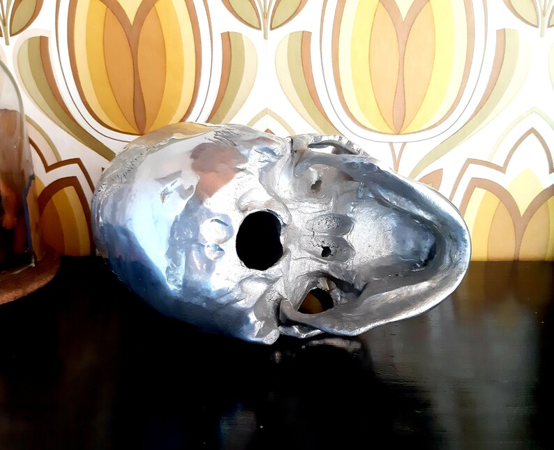 May include: A silver skull sculpture with a detailed design. The skull is hollow and has a large eye socket opening.