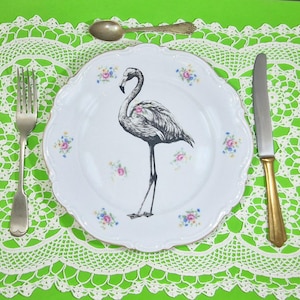 Pode incluir: Um prato de cerâmica branco com uma ilustração de flamingo, rodeado por detalhes florais. Um garfo, uma colher e uma faca prateados com cabo dourado estão dispostos sobre uma superfície verde com uma toalha de renda branca.