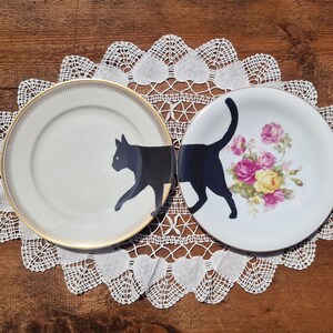 May include: Two round ceramic plates featuring a black cat graphic. The left plate has a gold rim and shows the front of a black cat. The right plate displays the rear of the same black cat with a cluster of pink and yellow roses beside it.