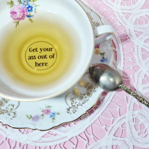 Puede incluir: Un juego de taza y platillo blancos con diseños florales y ribetes dorados, lleno de té. La base de la taza tiene el texto "Get your ass out of here". Una cuchara plateada descansa sobre un tapete de encaje rosa.