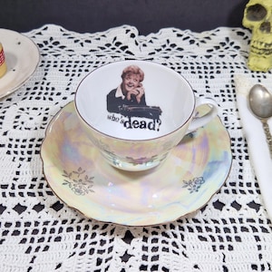 Può includere: Un set vintage di tazza e piattino da tè con finitura perlata. La tazza presenta un'immagine in bianco e nero di una donna con la scritta "who's dead?". Il piattino ha un bordo smerlato e decorazioni floreali. Un cucchiaio e un teschio sono sullo sfondo.