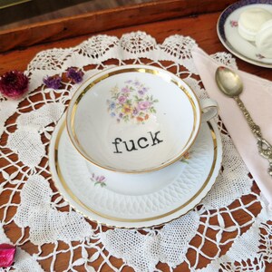 Può includere: Tazza e piattino bianchi con bordatura dorata e la parola "fuck" stampata in nero. L'interno della tazza presenta un motivo floreale. Il set è su un centrino di pizzo bianco, con un cucchiaio e dei macarons.