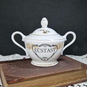 May include: White porcelain sugar bowl with lid, featuring gold accents and the word "ECSTASY" in a black, ornate frame. The bowl has two handles and a small knob on the lid. The design includes gold floral patterns.