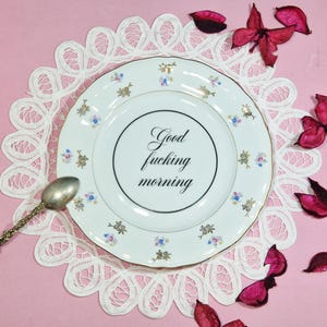 May include: White porcelain plate with gold trim and floral accents, featuring the text "Good fucking morning." The plate rests on a white lace doily, with a silver spoon and scattered pink petals. A unique and decorative item.